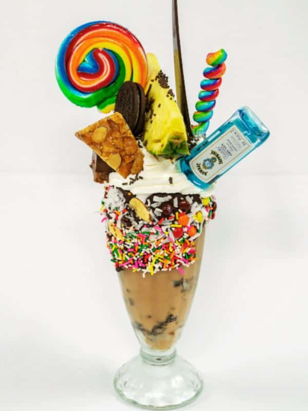 A decadent milkshake served on a Princess Cruises ship, garnished with colorful sprinkles, whipped cream, a rainbow lollipop, an Oreo cookie, a pineapple slice, a piece of almond brittle, a swirl lollipop, and a mini bottle of Bombay Sapphire gin.