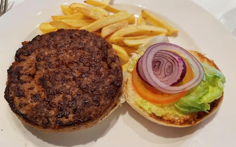 A plate of food on a Princess Cruises ship featuring a hamburger patty on one half of a bun, and the other half topped with lettuce, a slice of tomato, and red onion rings. The meal is accompanied by a side of French fries.