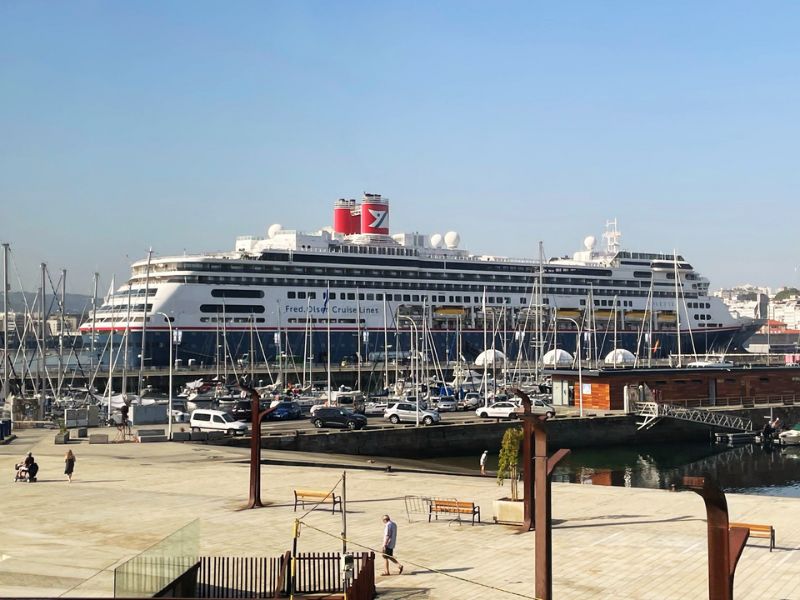 A Fred. Olsen Cruise Lines ship docked at the port of La Coruña, Spain. The vessel, characterized by its large size and distinctive red funnel with the Fred. Olsen logo, is moored at the harbor, surrounded by a bustling marina filled with smaller boats and yachts. The background features a clear blue sky and the urban landscape of La Coruña, providing a scenic view. The foreground of the photo shows a spacious, well-maintained promenade along the waterfront, where a few pedestrians are seen enjoying the sunny weather. This setting highlights the integration of cruise tourism within the coastal cityscape, offering visitors an accessible gateway to explore the region’s cultural and scenic attractions.