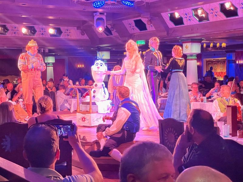 A vibrant live performance of a "Frozen" themed show aboard the Disney Wish cruise ship. The scene is lively with characters from the movie, including Elsa and Anna in their iconic dresses, and Olaf the snowman, engaging with the audience. A performer dressed as Kristoff, complete with traditional attire, is also visible. The setting is an ornately decorated dining area, illuminated by theatrical lighting that adds a magical glow to the performance. Guests are seated around, some capturing the moment on their devices, clearly enjoying the immersive entertainment experience unique to Disney cruises.