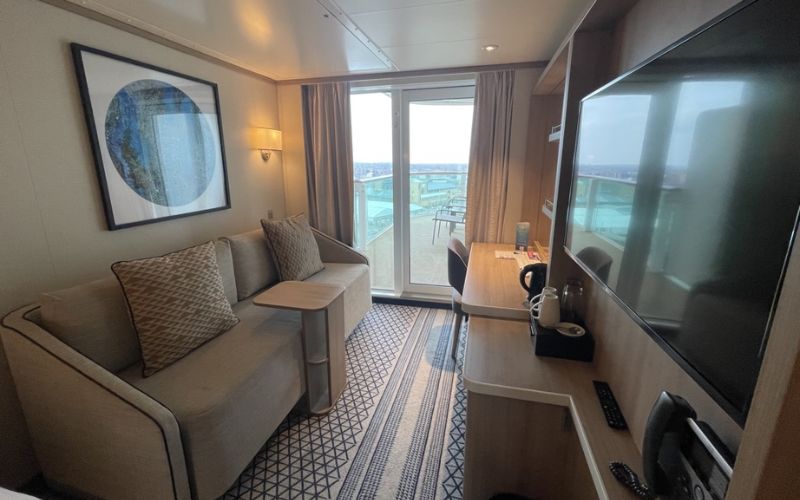 Inside a P&O Cruises Iona Balcony Cabin, a comfortable lounge area with a beige sofa and cushions, a wooden coffee table, and a large wall-mounted TV. The sliding glass door opens to a balcony with a table and chairs, offering a panoramic view of the sea.