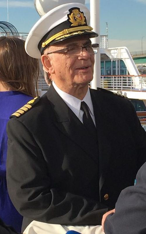 Close-up of an elderly gentleman wearing a naval officer's uniform with a white captain's hat and gold accents on the shoulders. He appears to be part of an event, with a hint of a blue top visible in the background along with parts of a cruise ship.