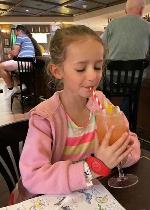 A young girl enjoying a fruity drink with a striped straw, dressed in a striped top and pink hoodie, seated in a casual dining setting with patrons in the background.