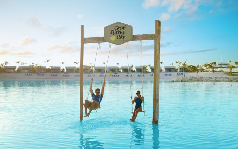 A man and woman are swinging on large wooden swings set in the shallow waters of Great Stirrup Cay. The swings are suspended from a wooden frame with a sign that reads "Great Stirrup Cay." The serene beach scene in the background features palm trees, cabanas, and a clear blue sky, creating a perfect tropical getaway ambiance.