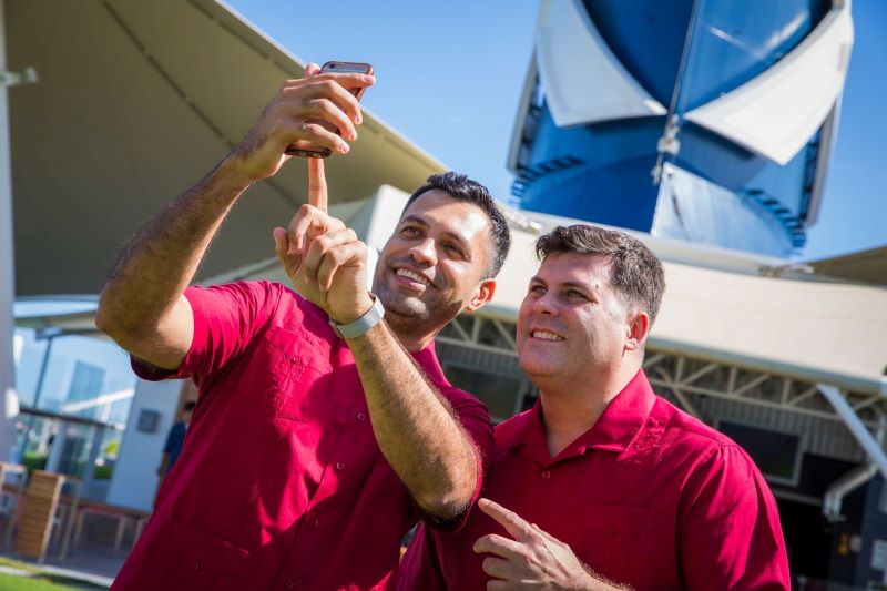 Two guests in matching red shirts are taking a selfie onboard a Celebrity Cruises ship, with the distinctive blue and white ship funnel in the background, capturing a joyful moment of their vacation.