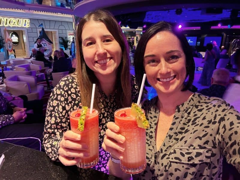 Two smiling guests are enjoying their vacation on a cruise ship, toasting with vibrant pink drinks in a lively lounge area, sharing a moment of relaxation and joy together.