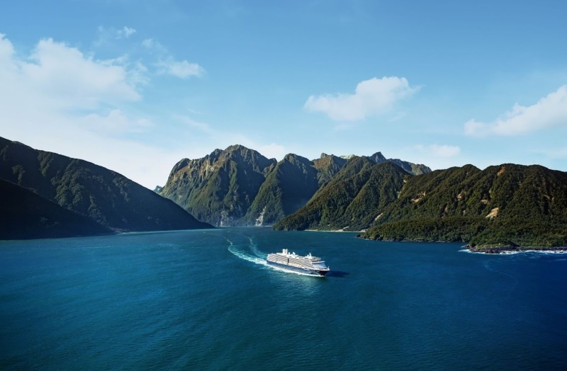 A Holland America Line cruise ship traversing the deep blue waters of a fjord surrounded by lush green mountains under a clear blue sky.
