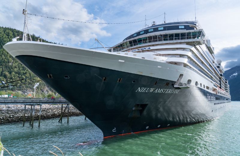 Holland America Line cruise ship, Nieuw Amsterdam, docked at the port of Skagway, Alaska. The large, impressive vessel, painted in dark blue with white superstructures, is set against the rugged and scenic backdrop typical of Alaskan ports. The photo captures the ship's sleek design and multiple decks, highlighting its capacity for luxury travel. The surrounding environment, with its mountainous terrain and clear waters, emphasizes the natural beauty and remote wilderness that attract visitors to this part of Alaska. This setting provides cruise passengers with a unique opportunity to explore the historical and picturesque town of Skagway, known for its role in the Klondike Gold Rush.
