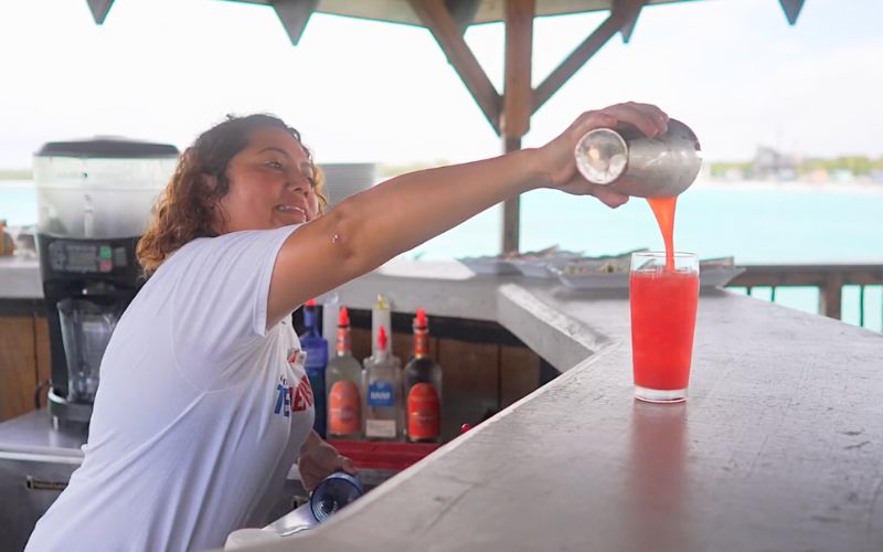 A bartender at Half Moon Cay pours a vibrant red cocktail with a smile, capturing the joyful spirit of the tropical getaway. The bar’s casual setting is highlighted by the clear turquoise waters in the background, inviting guests to relax and enjoy the island’s festive atmosphere.