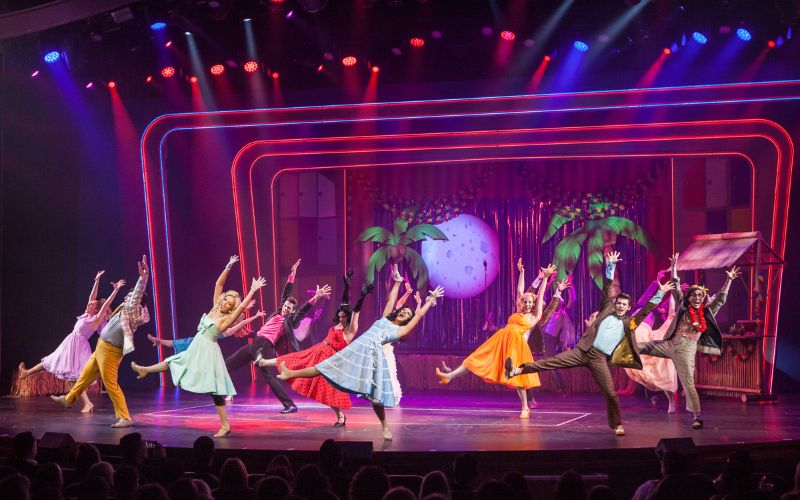 A vibrant scene from a Broadway-style show on Harmony of the Seas features a group of performers in colorful costumes energetically dancing on stage. The backdrop includes palm trees and a large decorative moon, with bright, multicolored stage lighting enhancing the lively atmosphere. The performers strike dynamic poses, engaging the audience with their enthusiastic performance.