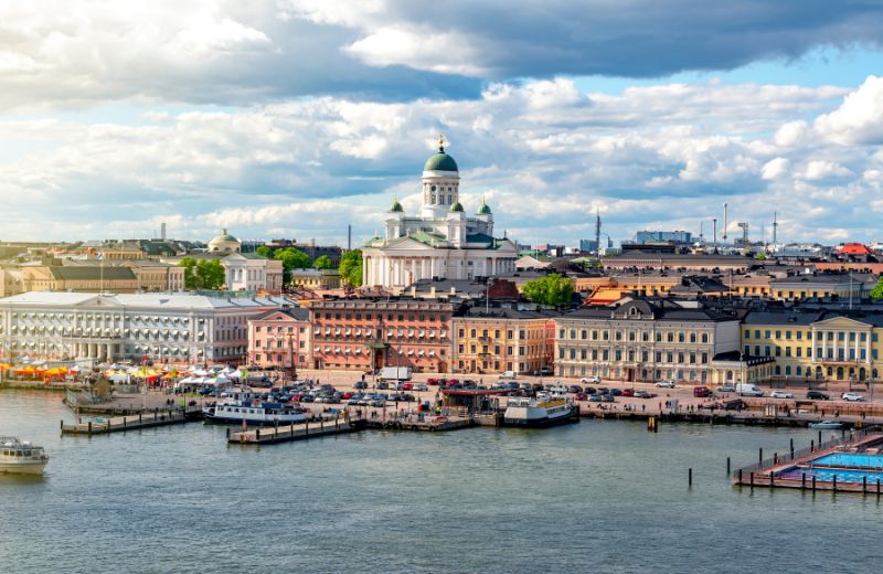 A breathtaking view of Helsinki, Finland, showcasing the iconic Helsinki Cathedral dominating the skyline with its green dome and white exterior. The cathedral is a focal point against the backdrop of Helsinki's cityscape, which includes classic European architecture in shades of pastel, bustling harbor activities, and modern infrastructure. The scene is alive with both marine and urban elements, with boats moored at the docks and people likely enjoying the waterfront. This panoramic view under a partly cloudy sky vividly illustrates the blend of historical significance and contemporary lifestyle that Helsinki offers.