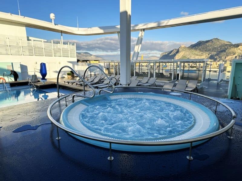 A bubbling hot tub on the deck of a cruise ship awaits passengers, with a view of tranquil blue skies and a picturesque mountain in the distance, offering a relaxing retreat amidst the beauty of the cruising route.