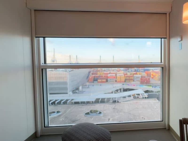 View from the 'Infinite Balcony' aboard Royal Caribbean's 'Star of the Seas,' showing a bustling port scene. The large window frames a view of colorful cargo containers and the port infrastructure, blending the indoor comfort with the industrial exterior. A subtle, wicker chair is partially visible, enhancing the room's connection to the outdoor scenery.