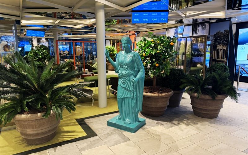 Interior of the Costa Smeralda featuring a teal replica of the Statue of Liberty, surrounded by lush green potted plants, under a ceiling of colorful destination signs and digital screens.