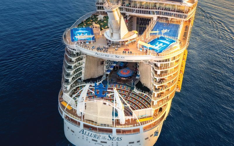 Top-down view of the 'Allure of the Seas' cruise ship's aft, showcasing the tiered deck structure with a sports court, pools, and sun loungers. The ship's name is prominently displayed on the stern, against the calm blue ocean backdrop.