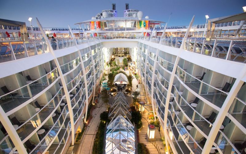 An interior view of a cruise ship's central promenade at dusk, featuring a multi-level design with private balconies facing the indoor park filled with lush greenery and a glass-roofed atrium, festively adorned with international flags.