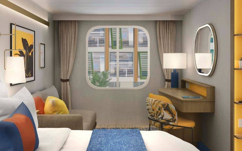Interior view of another stateroom aboard Royal Caribbean's 'Star of the Seas,' showcasing modern amenities and décor. The room features a plush bed with blue and white bedding, colorful accent pillows, a beige sofa, and a small work desk with an oval mirror. A large porthole-style window provides a view of the ship's exterior passageways, adding natural light to the space.