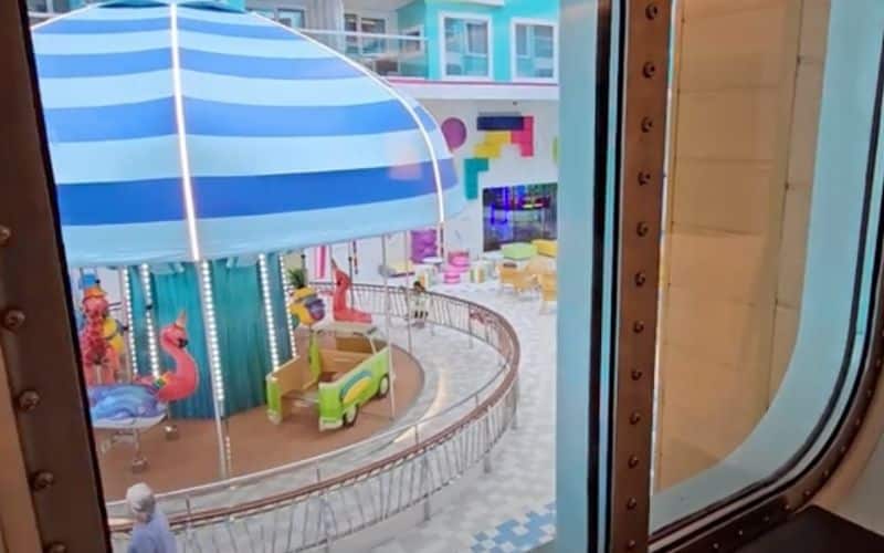 View from the window of an interior stateroom on Royal Caribbean's 'Star of the Seas,' overlooking the ship's colorful and lively Surfside neighborhood. The window frames a bustling children's area featuring a whimsical carousel under a large blue and white striped umbrella, surrounded by vibrant playground equipment and seating areas.