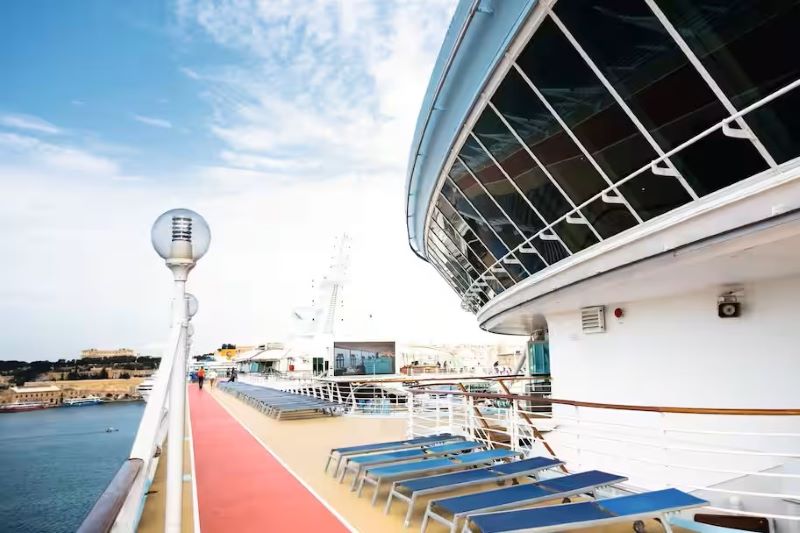 This image offers a view from the deck of a cruise ship, featuring a series of blue lounge chairs lined up along a red walkway. The photo is taken looking towards a distinctive glass-enclosed structure on the ship, possibly an observation lounge or restaurant, with the coastal cityscape in the background under a clear sky. This setting emphasizes the leisure and luxury aspects of cruise travel, highlighting spaces designed for relaxation and enjoyment of the seascape.