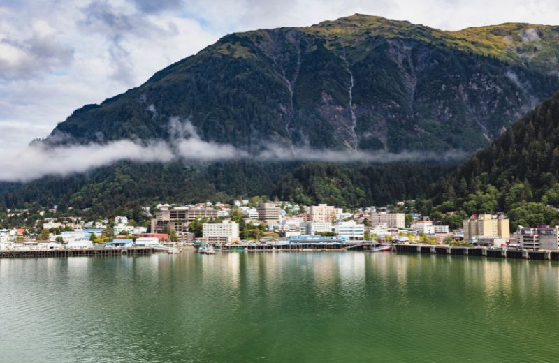 A striking view of Juneau, the capital city of Alaska, set against a dramatic natural backdrop. Nestled at the water's edge, the city's buildings are modest in height, reflecting the small-scale urban development typical of this remote area. Behind the city, a lush, green mountain rises steeply, shrouded partially by low-hanging clouds that add a mystic quality to the landscape. The calm, greenish waters in the foreground reflect the tranquility of this picturesque setting, emphasizing Juneau's unique blend of urban life and wild, natural beauty. This view highlights the city's isolated and scenic location, surrounded by the vast wilderness that is characteristic of Alaska.