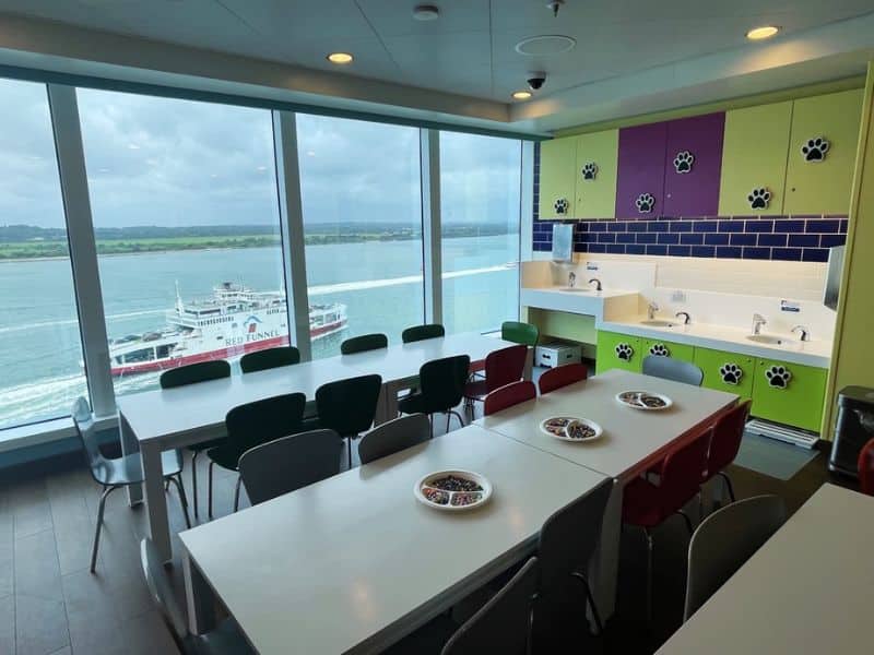 A kids' club area on a Princess cruise ship featuring tables and chairs set up for activities, with bowls of small toys or candies on the tables. The room has colorful cabinets with paw print decorations, a row of sinks, and large windows offering a view of the ocean and a passing Red Funnel ferry.