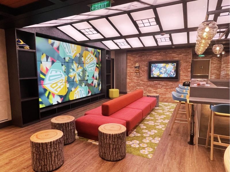 A modern and stylish kids' club area aboard a Disney cruise ship. The space features urban-inspired decor with exposed brick walls and a chic skylight ceiling design, creating a bright and airy atmosphere. There's a large, vibrant geometric pattern displayed on a massive screen, adding a dynamic and colorful element to the room. In the foreground, a comfortable red sectional sofa and tree stump stools offer plenty of seating for kids to relax or watch shows. The right side of the room includes a bar area with high stools, ideal for activities or dining. This setting provides a hip, lounge-like environment that appeals to both younger guests and teenagers.