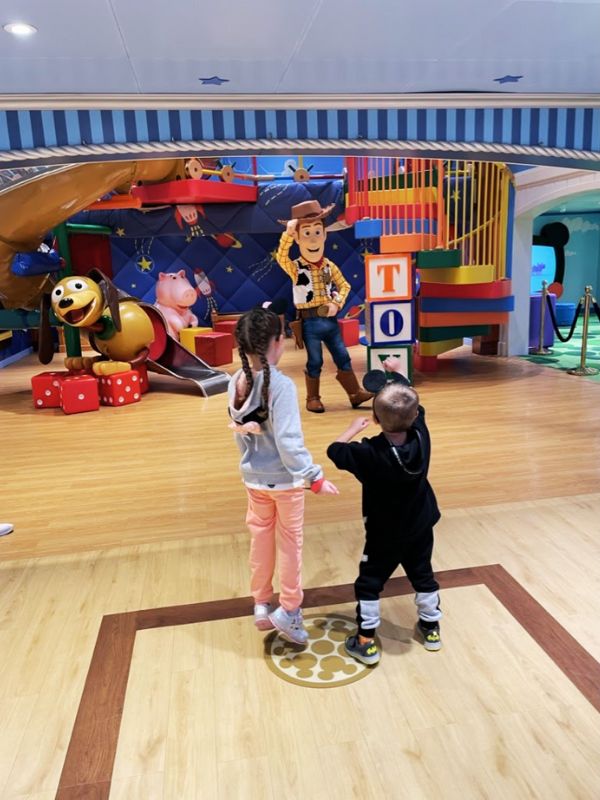 A delightful scene inside a kids' club on a Disney cruise ship, designed with a vibrant, Toy Story-themed play area. Two young children, a girl and a boy, are interacting with a life-sized figure of Woody, the beloved cowboy character from the Toy Story series. Woody stands with open arms, welcoming the kids who are excitedly approaching him. The play area is colorful and inviting, featuring large building blocks, a Slinky Dog slide, and other Toy Story decorations, creating an immersive environment for children to play and meet their favorite characters.