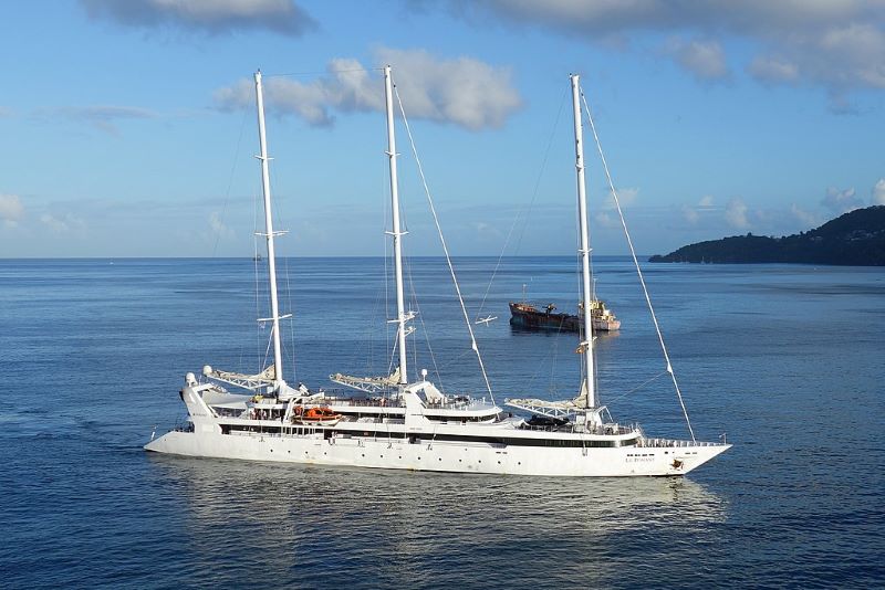The sailing yacht Le Ponant, a sleek white vessel with three prominent masts, each equipped with furled sails. The yacht is seen cruising on tranquil blue waters with a scenic coastline and hills visible in the background. There is a freighter ship in the distance, providing a contrast to the elegant and leisurely vibe of Le Ponant under a clear sky.