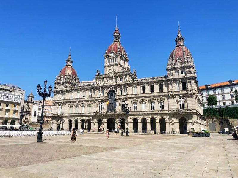 The grand City Hall in La Coruña, Spain, located in the Maria Pita Square. This iconic building is renowned for its ornate architectural design, featuring elaborate stone carvings, intricate facades, and two prominent domed towers. The clear blue sky and the spacious square in front provide a perfect backdrop that highlights the building's historic elegance. The area is mostly devoid of crowds, offering a calm and open view of this majestic city landmark, making it an inviting scene for visitors and locals alike to enjoy the cultural and architectural heritage of La Coruña.