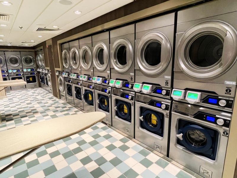 Laundry area on a Disney cruise ship, designed for guest convenience. The room is equipped with multiple commercial-grade washing machines and dryers, neatly aligned and organized. Each machine has a digital interface indicating its status, enhancing user friendliness. The floor is tiled with a light and dark blue checkerboard pattern, and there is a large ironing board in the foreground, indicating the facilities are well-prepared for all laundry needs. The overall clean and modern design of the laundry room ensures a pleasant experience for guests needing to wash clothes during their cruise.