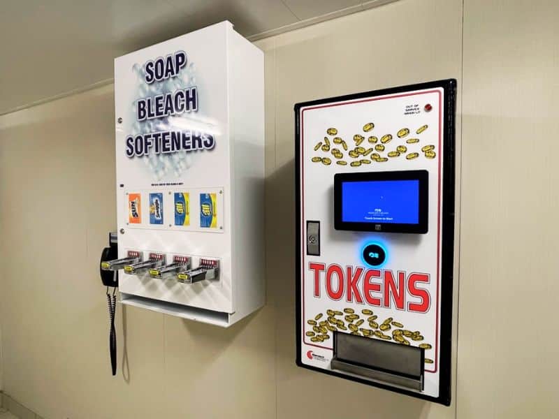 A laundry area on a Princess cruise ship featuring a vending machine for laundry supplies such as soap, bleach, and softeners. Next to it is a token machine with a digital screen, used for purchasing tokens for the laundry machines. Both machines are mounted on a beige wall.