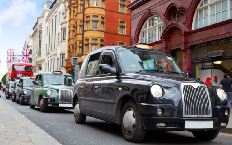 A line of iconic black London taxis is seen on Oxford Street, with the characteristic red-brick architecture of the area in the background. British flags are displayed along the street, and a red double-decker bus is visible, enhancing the quintessential London scene.