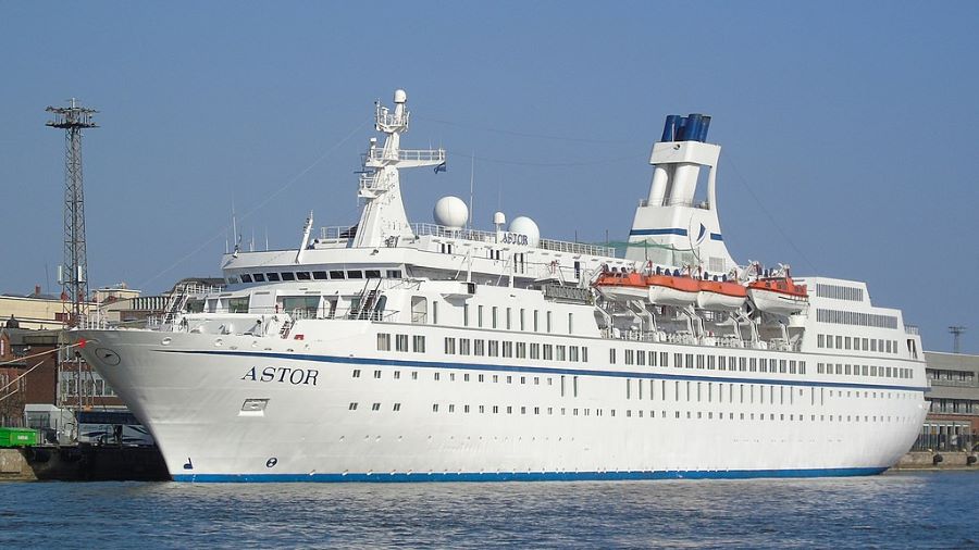 The cruise ship MS Astor docked at a port, with clear blue skies above and an urban setting in the background. The ship's white exterior is accented with a blue stripe running along the lower deck and a distinctive blue and white funnel. Lifeboats are visibly mounted on both sides, and various antennas and radars can be seen atop the bridge, highlighting the vessel's equipped and ready-for-sail status.