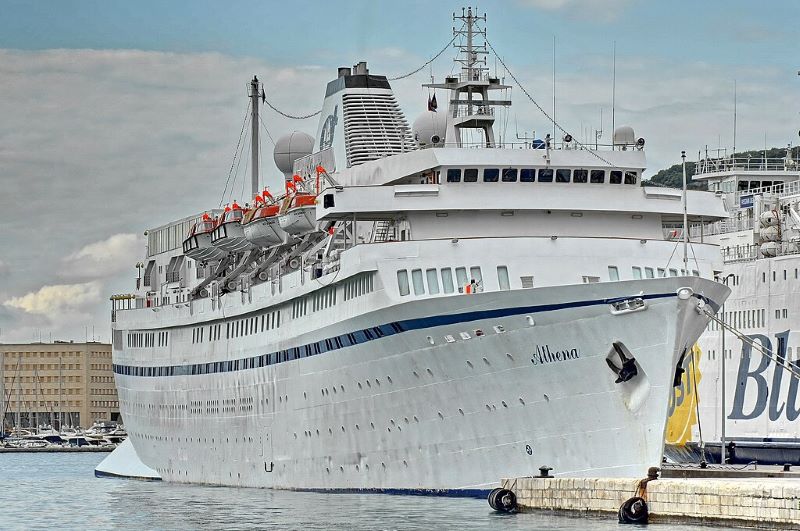 The MS Athena docked at a port under a cloudy sky, capturing a dramatic and detailed view of the cruise ship. The ship's white and blue paint highlights its elegant design, featuring multiple decks adorned with lifeboats and advanced navigational equipment on the bridge. The bustling port setting and industrial backdrop contrast with the ship's leisurely purpose, emphasizing its readiness for voyages.