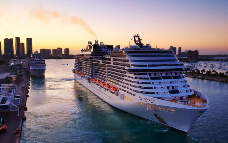The MSC Meraviglia cruise ship departs from a port at sunset, its decks lined with lifeboats and gleaming under the warm sky, juxtaposed against a backdrop of urban high-rises, embodying the elegance of oceanic travel.