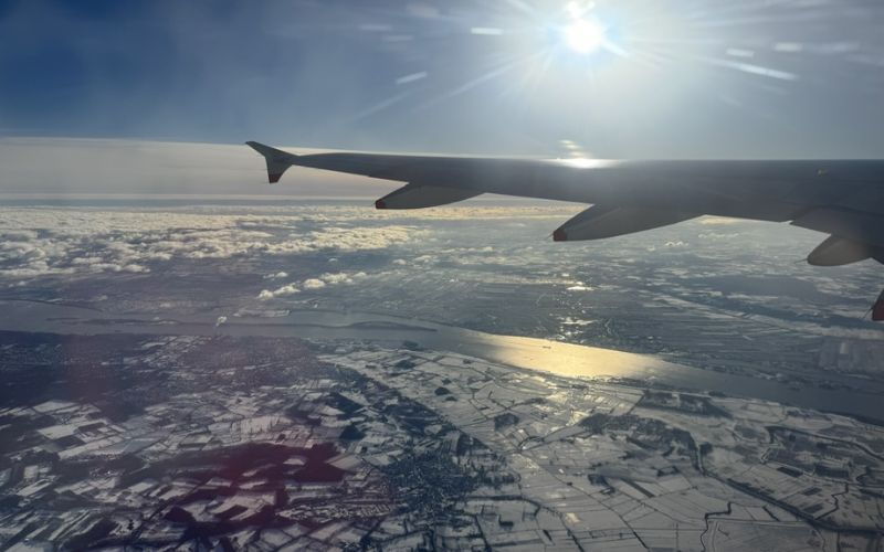 A view from an airplane window shows the wing of the aircraft against a backdrop of a clear blue sky and the sun shining brightly. Below, a landscape of fields and rivers is partially covered in snow, with clouds scattered across the horizon, creating a serene and picturesque aerial scene.