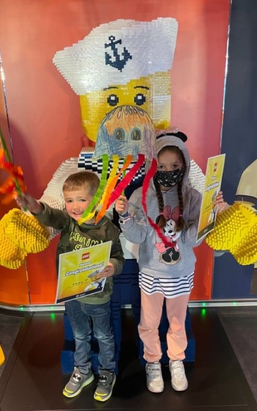 Two children pose happily with certificates in front of a large LEGO sailor statue at an MSC Cruise Kids' Club. The boy on the left holds colorful streamers and a "Congratulations" certificate, while the girl on the right, wearing a Minnie Mouse hoodie and a face mask, holds a similar certificate and streamers. The background features bright colors and playful decorations, enhancing the fun atmosphere.