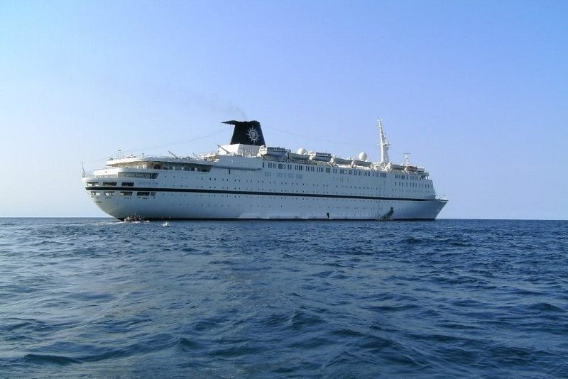 The MSC Melody cruise ship sailing on open blue waters. The ship, predominantly white with distinctive black and blue funnel bearing the MSC logo, spans across the frame displaying its length and multiple decks lined with windows. The tranquil sea and clear skies provide a serene setting, enhancing the ship’s graceful voyage.