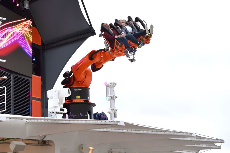 Passengers experiencing the thrilling Robotron ride on the MSC Seascape cruise ship, a robotic arm lifting a secured roller coaster-style seat high above the deck with a dynamic LED screen display in the background.