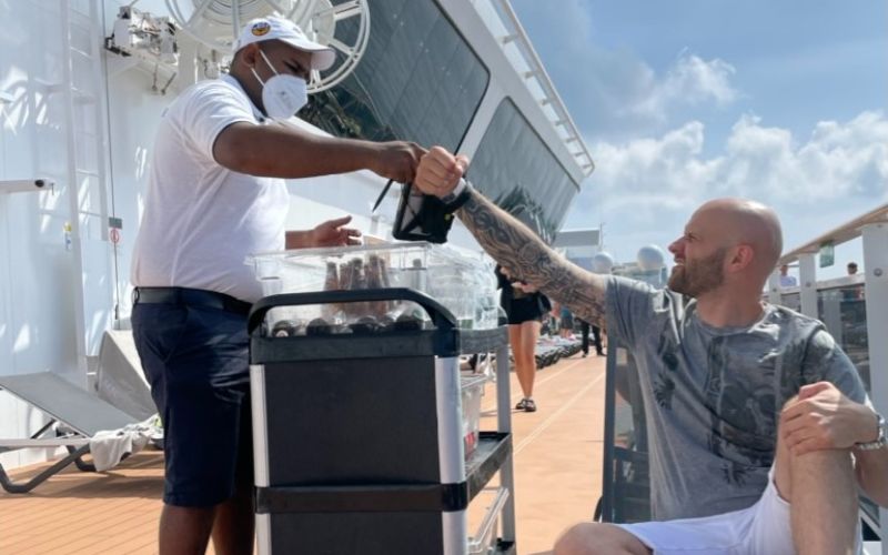A crew member wearing a mask and uniform serves a guest seated on the deck of a cruise ship. The guest, a bald man with tattoos, is receiving a drink from a cart filled with various beverages. The scene takes place under a bright, sunny sky, with the ship's sleek exterior and other passengers in the background.
