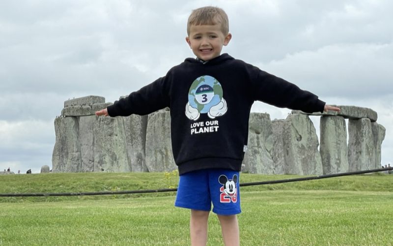 My son on an MSC shore excursion. He smiles with his arms outstretched, standing in front of the iconic Stonehenge monument. He is wearing a black hoodie with a "Love Our Planet" design and blue shorts featuring a Mickey Mouse patch. The grassy field and ancient stones create a historic and picturesque backdrop under a cloudy sky.