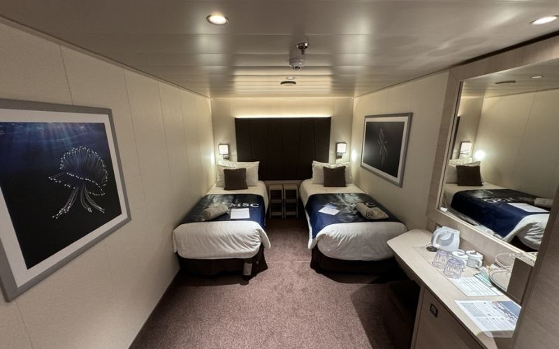 A cosy cabin on MSC Euribia cruise ship features two single beds with white linens and blue throws, separated by a nightstand. The room is decorated with framed marine-themed artwork and includes a vanity desk with a mirror and various amenities. Soft lighting creates a warm and inviting atmosphere.