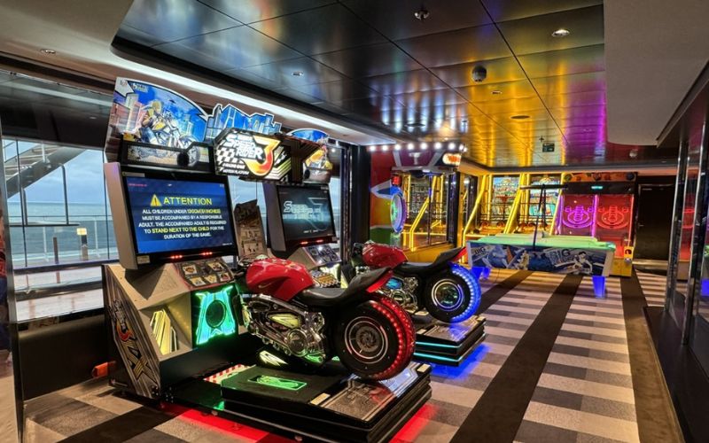 An arcade on a cruise ship features various games, including motorcycle racing simulators, air hockey, and other interactive attractions. The area is brightly lit with colorful neon lights, creating an exciting and playful atmosphere. Large windows offer a view of the sea, adding to the unique experience of gaming at sea.