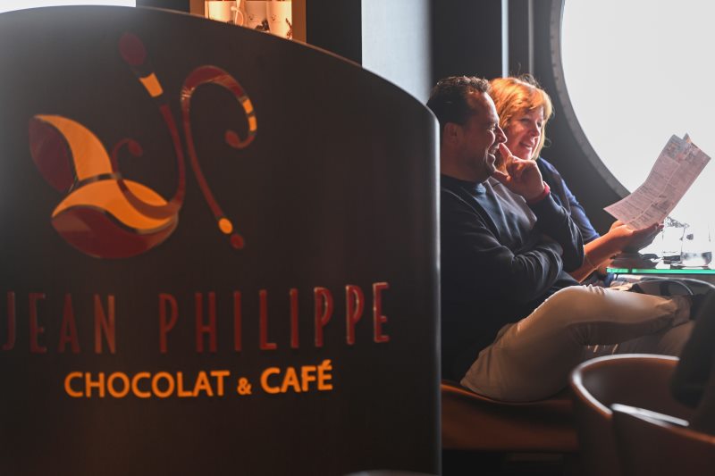A couple sits inside the Jean Philippe Chocolat & Café on an MSC cruise ship, reading the MSC daily program. The man and woman are engaged in conversation and appear to be planning their day. The café sign is prominently displayed in the foreground, while a large round window in the background offers a glimpse of the outside.