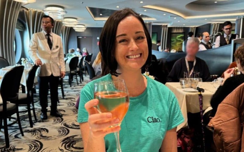 Cruise Mummy raises a glass of amber-colored beverage in the Main Dining Room on an MSC cruise ship, with elegant décor and attentive staff in the background, capturing a moment of joy and fine dining on a luxury voyage.