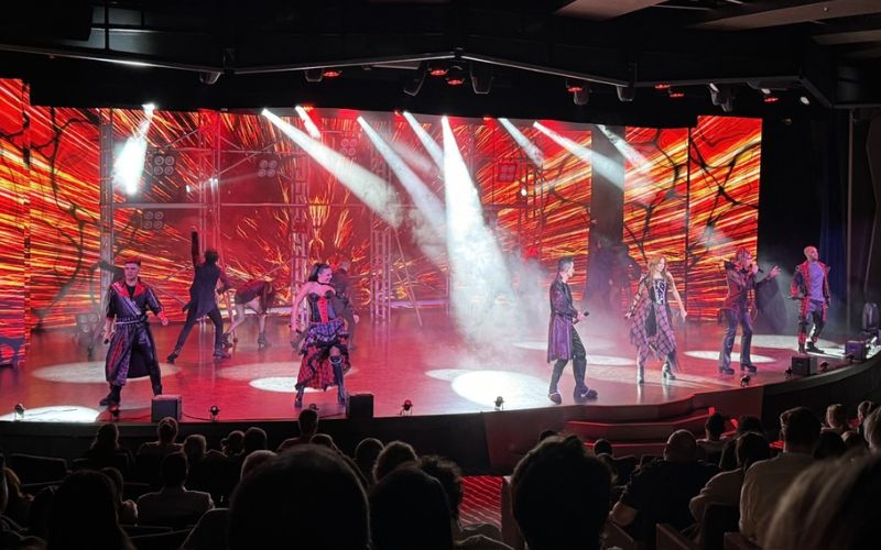 A vibrant stage performance on a cruise ship features a group of performers dressed in futuristic costumes under dynamic lighting. The background is illuminated with striking red and orange patterns, creating an energetic atmosphere. The audience is seated, watching the engaging and lively show.