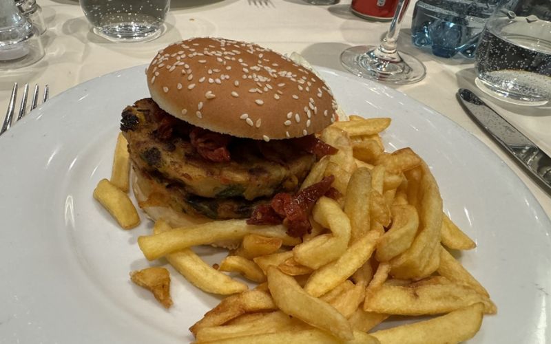 A plate of food featuring a sesame seed bun burger with a vegetable patty and sundried tomatoes, accompanied by a generous portion of golden French fries. The meal is set on a white tablecloth with silverware and a glass of sparkling water visible in the background.