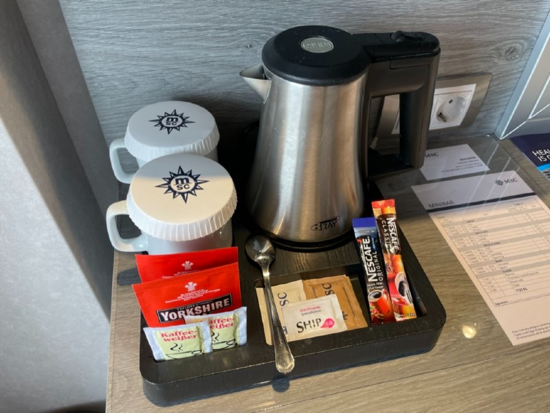 A neatly arranged tea and coffee station in a cabin on an MSC cruise ship includes a stainless steel electric kettle, two branded mugs with covers, a spoon, and a variety of tea and coffee sachets. The station is set on a wooden countertop with power outlets and cruise information paperwork nearby.