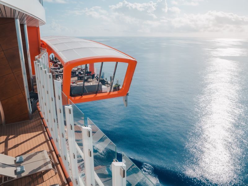 The Magic Carpet on a Celebrity Cruise ship, an innovative cantilevered floating platform with orange accents, offers guests a unique vantage point as it extends out from the ship, overlooking the tranquil blue ocean waters.