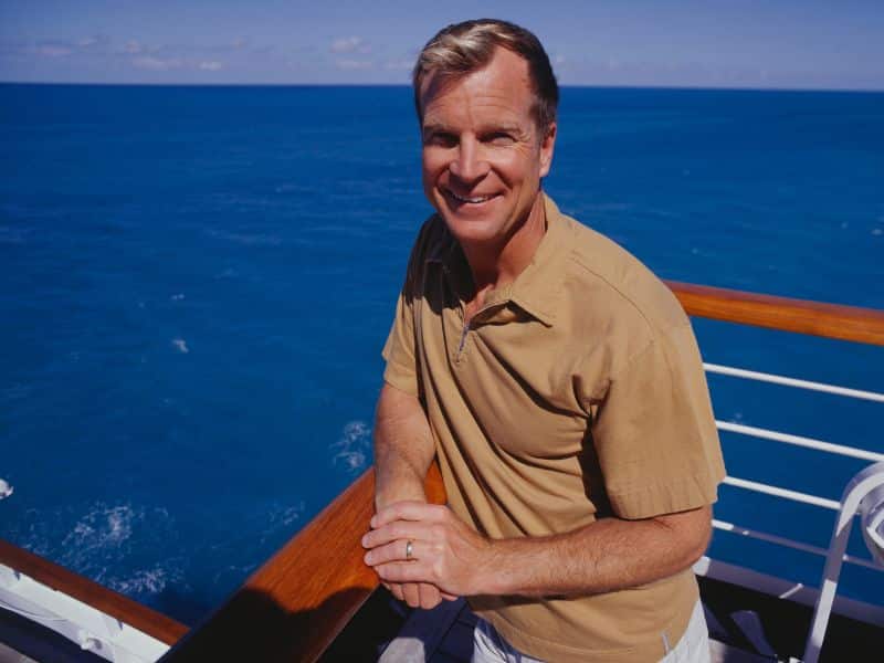 A smiling man leaning on the railing of a cruise ship deck with the deep blue sea in the background under a clear sky.
